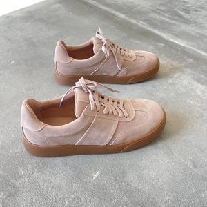 May include: A pair of light pink suede sneakers with a brown rubber sole. The sneakers have a lace-up closure and a simple design.