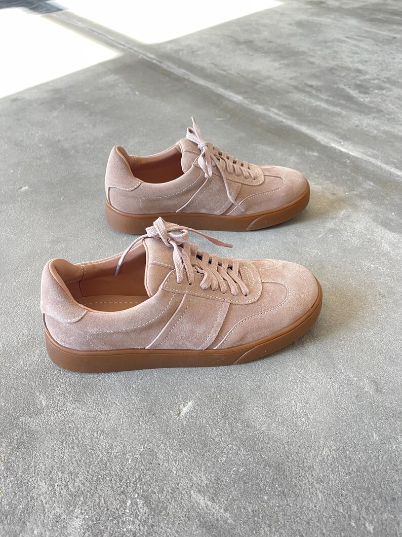 May include: A pair of light pink suede sneakers with a brown rubber sole. The sneakers have a lace-up closure and a simple design.