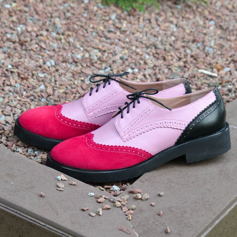 Pu&ograve; includere: Un paio di scarpe in pelle rosa e rosse con dettagli neri e lacci. Le scarpe hanno un design brogue e un tacco spesso.