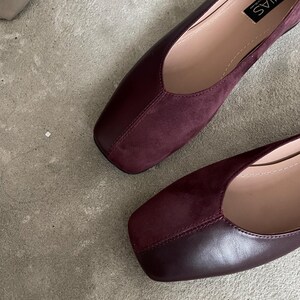 May include: A pair of burgundy leather and suede square-toe flats. The shoes have a simple design and are made of high-quality materials. The brand name "MIAS" is printed on the inside of one shoe.