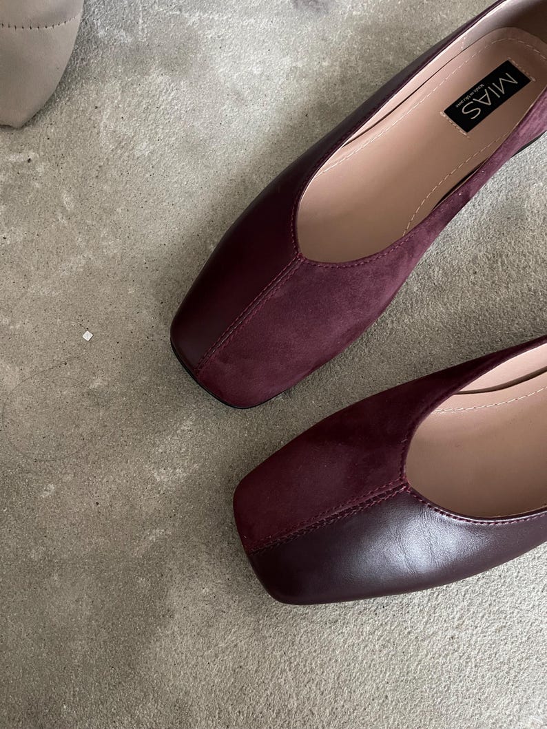 May include: A pair of burgundy leather and suede square-toe flats. The shoes have a simple design and are made of high-quality materials. The brand name "MIAS" is printed on the inside of one shoe.
