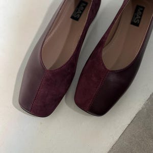 May include: A pair of burgundy suede and leather flats with a square toe. The shoes have a simple design and are labeled "MAS" on the inside.
