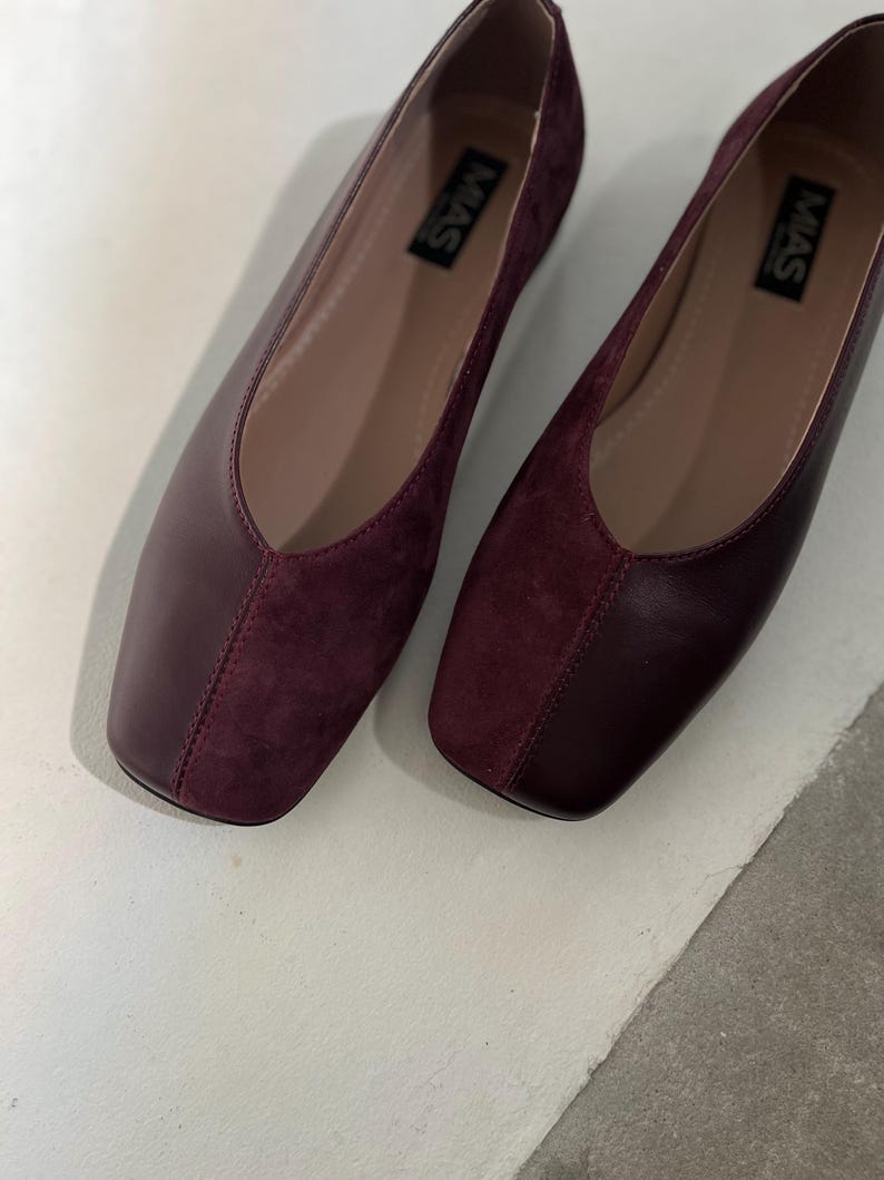 May include: A pair of burgundy suede and leather flats with a square toe. The shoes have a simple design and are labeled "MAS" on the inside.