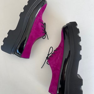 May include: A pair of fuchsia suede platform shoes with black patent leather accents and black laces. The shoes have a chunky platform sole.