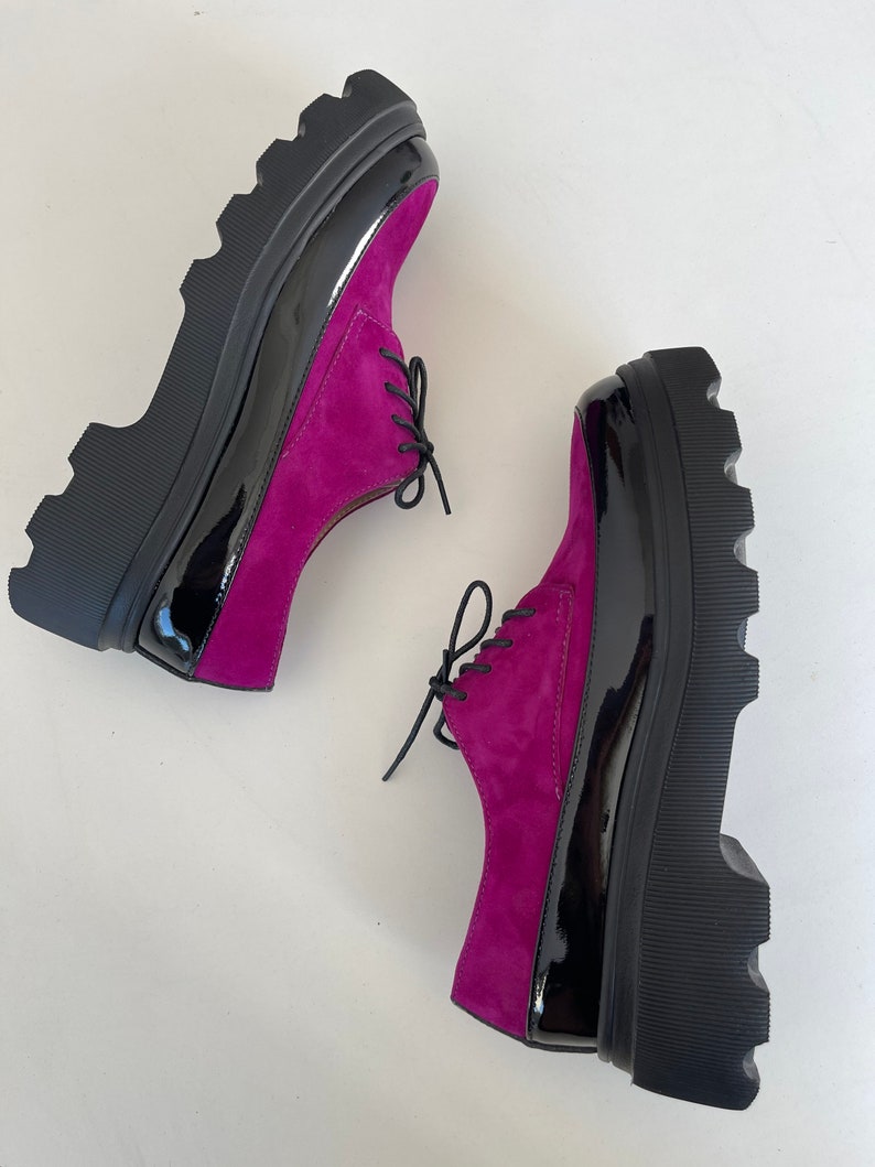 May include: A pair of fuchsia suede platform shoes with black patent leather accents and black laces. The shoes have a chunky platform sole.