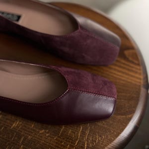 May include: A pair of burgundy leather flats with a suede toe cap. The shoes have a square toe and a low heel.