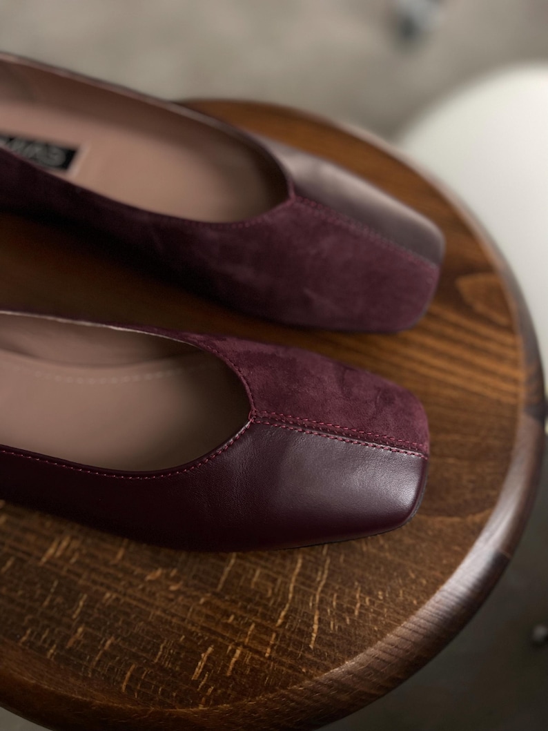 May include: A pair of burgundy leather flats with a suede toe cap. The shoes have a square toe and a low heel.