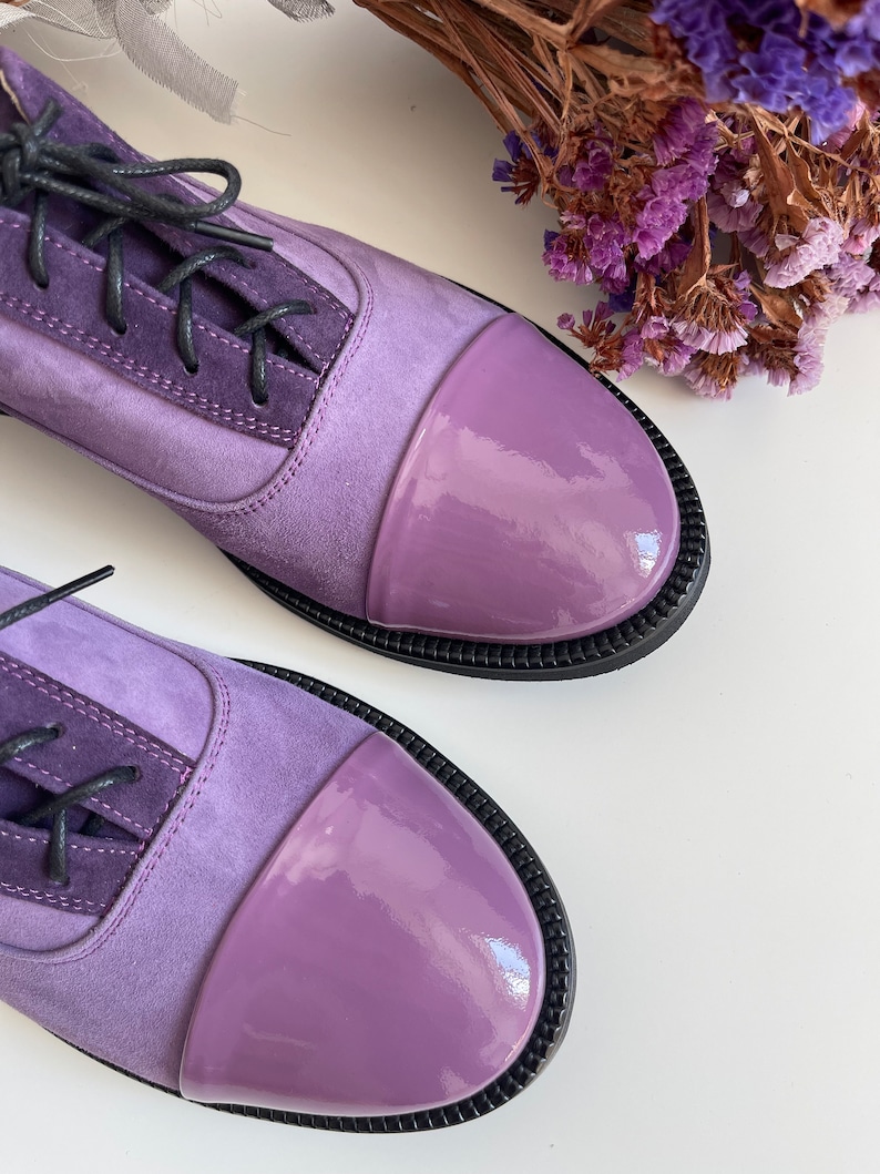 May include: A pair of purple suede and patent leather lace-up shoes with black laces. The shoes have a rounded toe and a black rubber sole.