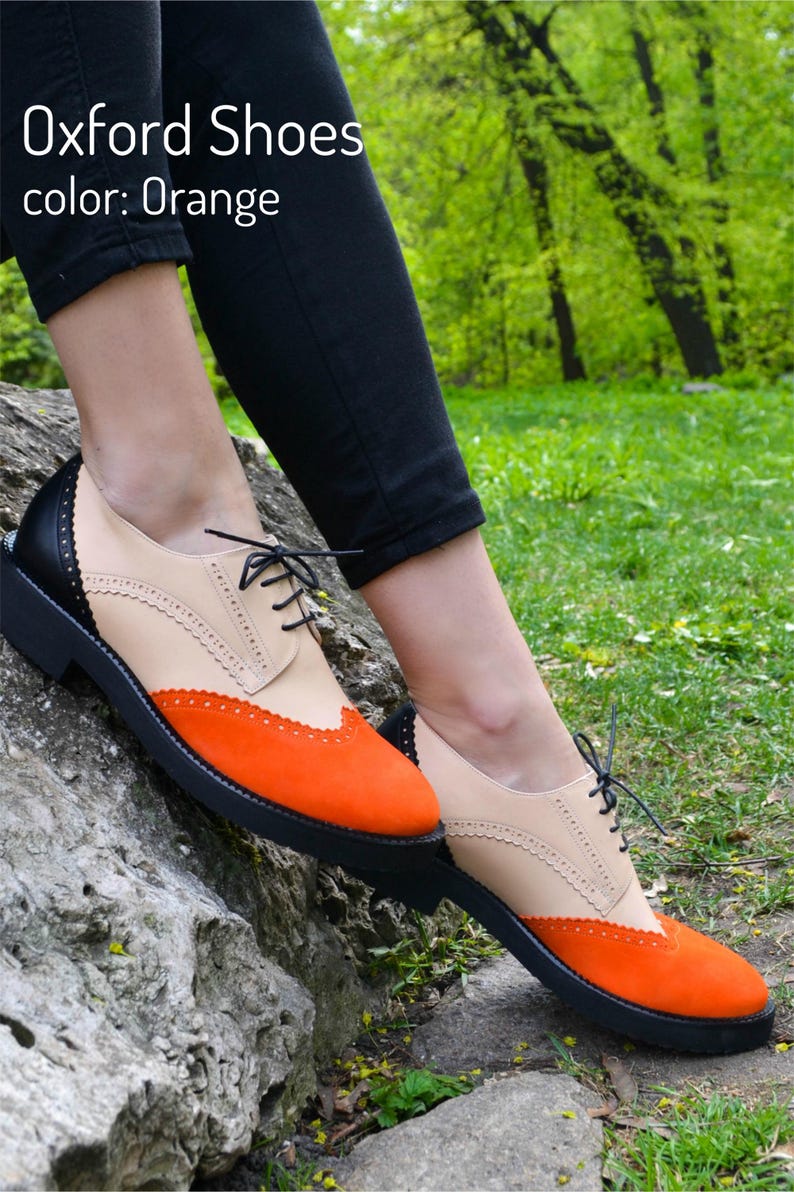 Pu&ograve; includere: Un paio di scarpe oxford arancioni e beige con lacci neri. Le scarpe hanno un design brogue e sono appoggiate su una roccia.