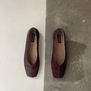 May include: A pair of burgundy flats with a square toe design. The shoes have a smooth leather upper and a suede vamp. The brand name "MIAS" is printed on the insole. The shoes are displayed on a two-tone background of white and gray.