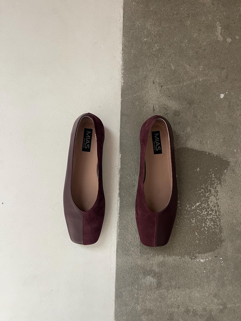 May include: A pair of burgundy flats with a square toe design. The shoes have a smooth leather upper and a suede vamp. The brand name "MIAS" is printed on the insole. The shoes are displayed on a two-tone background of white and gray.