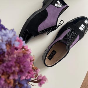 May include: A pair of black and purple leather oxfords with lace-up fastenings. The shoes have a black patent leather toe cap and a purple suede upper. The brand name "MIAS" is visible on the insole of one shoe.