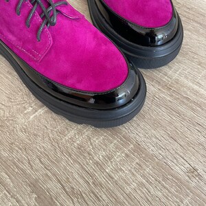 May include: A pair of fuchsia suede shoes with black patent leather trim and black laces. The shoes have a chunky platform sole.
