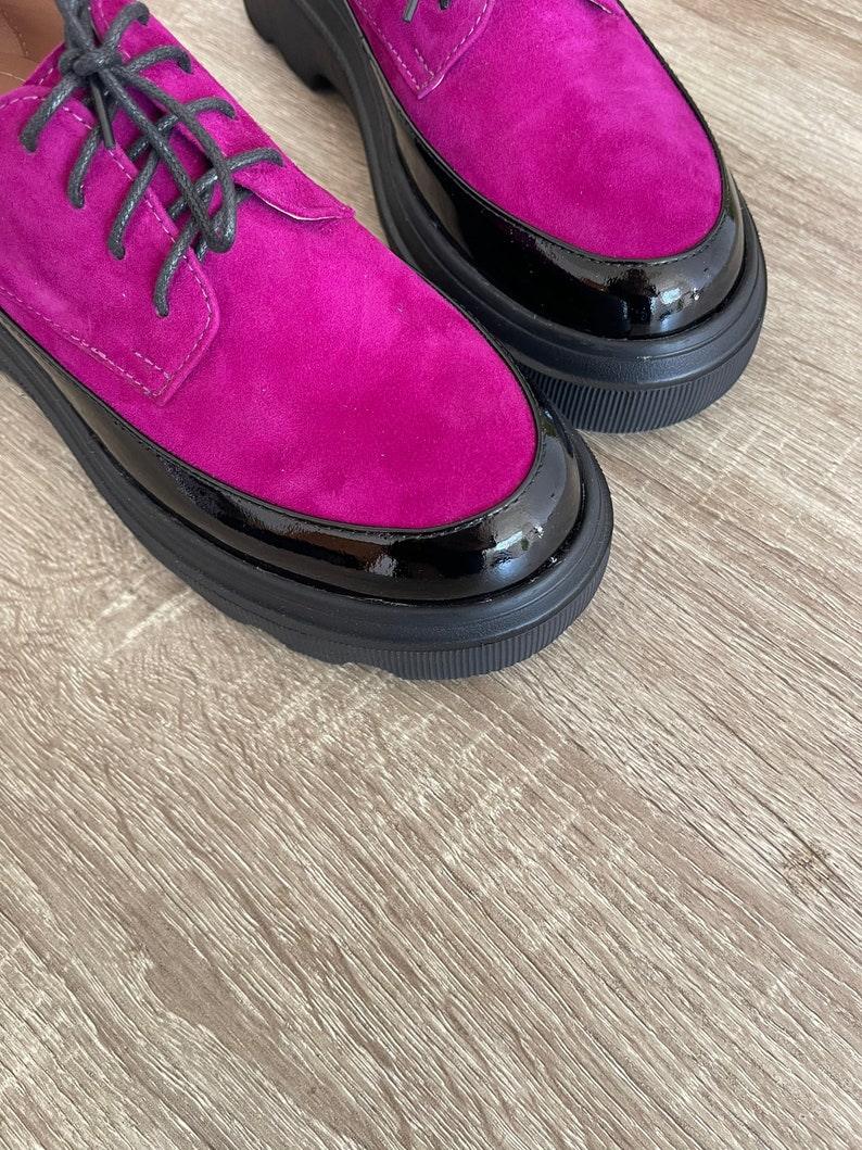 May include: A pair of fuchsia suede shoes with black patent leather trim and black laces. The shoes have a chunky platform sole.