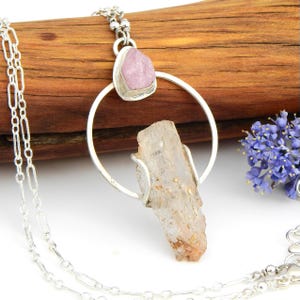Könnte beinhalten: Eine silberne Halskette mit einem großen, klaren Kristallanhänger und einem kleineren rosa Stein. Der Anhänger hängt an einer Silberkette. Die Halskette wird vor einem Holzgrund präsentiert.