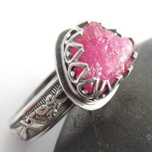 Può includere: Un anello d'argento con una pietra preziosa rosa grezza incastonata in una gabbia d'argento decorativa. La fascia dell'anello è decorata con un motivo floreale.