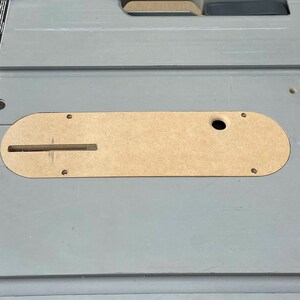 Pu&ograve; includere: Una sega da tavolo grigia con un inserto color beige e un metro a nastro giallo. La sega ha un telaio in metallo e diversi bulloni. Un interruttore di sicurezza arancione &egrave; visibile in basso.