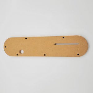 Puede incluir: Una plantilla de madera marr&oacute;n claro con varios agujeros y una ranura en el centro. La plantilla est&aacute; dise&ntilde;ada para proyectos de carpinter&iacute;a.