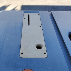 May include: A close-up of a gray table saw with a light brown insert. The insert has a slot and a hole. There is a small, round, orange sticker on the table.