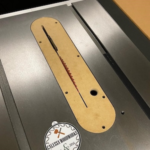 Puede incluir: Primer plano de una sierra de mesa con un inserto de madera y una hoja de sierra. La sierra de mesa tiene una superficie de metal plateado y un borde negro. Una pegatina con el texto "Calyssa Woodworks Est. 2016" est&aacute; en la sierra de mesa.