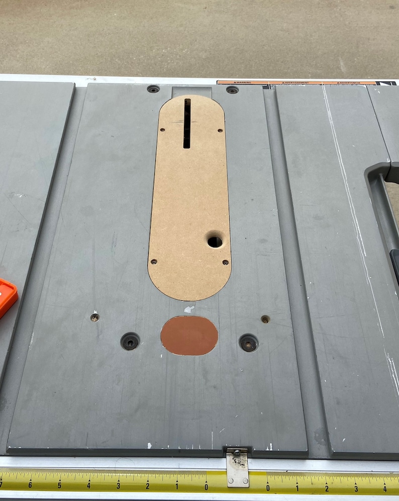 Pu&ograve; includere: Una sega da banco grigia con un inserto color beige e un inserto ovale marrone. La sega ha un righello di metallo lungo il bordo inferiore. La sega &egrave; progettata per la lavorazione del legno e il taglio di materiali.