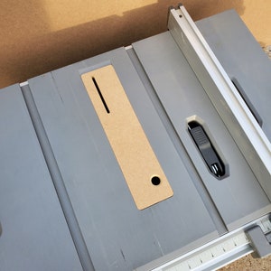 Op de afbeelding: Een grijze tafelzaag met een houten inzetstuk en een metalen hek. De tafelzaag heeft een liniaal die in inches is gemarkeerd.