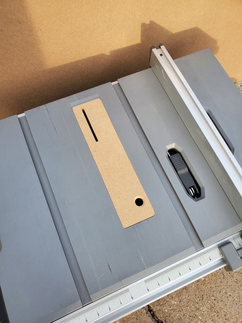 Op de afbeelding: Een grijze tafelzaag met een houten inzetstuk en een metalen hek. De tafelzaag heeft een liniaal die in inches is gemarkeerd.