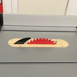 Puede incluir: Una sierra de mesa gris con una hoja de sierra roja y negra. La hoja est&aacute; parcialmente insertada en una abertura de madera en la mesa.