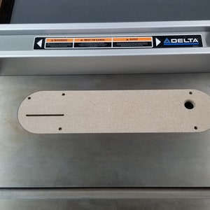 May include: A close-up of a Delta table saw with a router lift insert. The insert is made of a light brown material and has a slot for a router bit. The table saw is made of metal and has a silver finish.