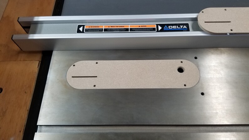 May include: A close-up of a Delta table saw with a router lift insert. The insert is made of a light brown material and has a slot for a router bit. The table saw is made of metal and has a silver finish.