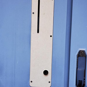May include: A close-up of a light blue surface with a rectangular cutout. The cutout is filled with a beige panel with a slot and a round hole. The panel is surrounded by black screws.