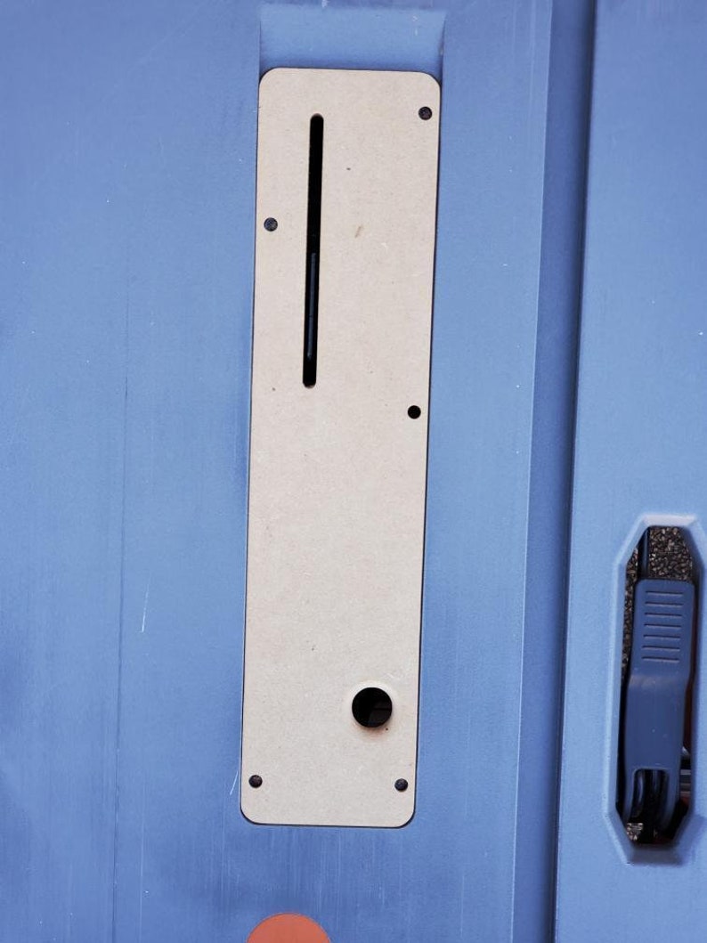 May include: A close-up of a light blue surface with a rectangular cutout. The cutout is filled with a beige panel with a slot and a round hole. The panel is surrounded by black screws.