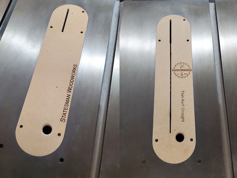 May include: Two beige wooden table saw inserts with a slot cut out in the center. The insert on the left has the text "STATESMAN WOODWORKS" engraved on it. The insert on the right has the text "Thin Kerf Straight" and a logo that says "CALYPSA WOODWORKS" engraved on it.