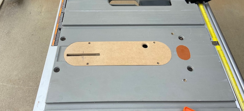 Pu&ograve; includere: Una sega da tavolo grigia con un inserto color beige e un metro a nastro giallo. La sega ha un telaio in metallo e diversi bulloni. Un interruttore di sicurezza arancione &egrave; visibile in basso.