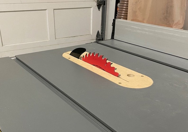 Puede incluir: Primer plano de una sierra de mesa con una hoja de sierra roja. La hoja es parcialmente visible a trav&eacute;s de una abertura de madera en la mesa.