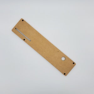 May include: A light brown wooden panel with a rectangular cutout and a round hole. The panel has four small black dots on the edges.