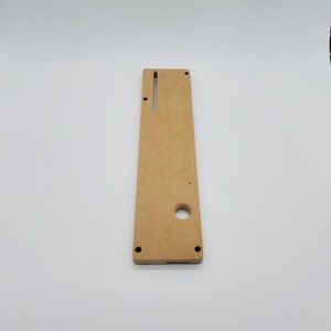 May include: A light brown wooden panel with a rectangular cutout and a round hole. The panel has two small black dots on the bottom edge.