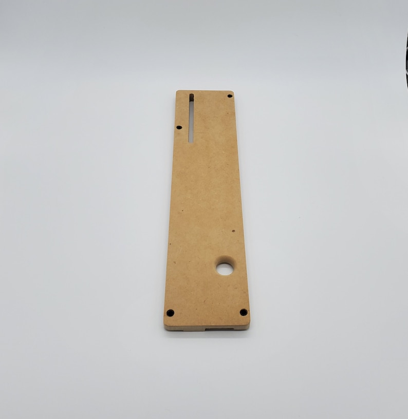 May include: A light brown wooden panel with a rectangular cutout and a round hole. The panel has two small black dots on the bottom edge.