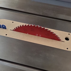 May include: A red saw blade with the number "100" printed on it is partially visible in a wooden slot on a metal table saw. The blade is surrounded by a wooden insert with a hole in the center. Two blue plastic pieces are visible on the left side of the insert.