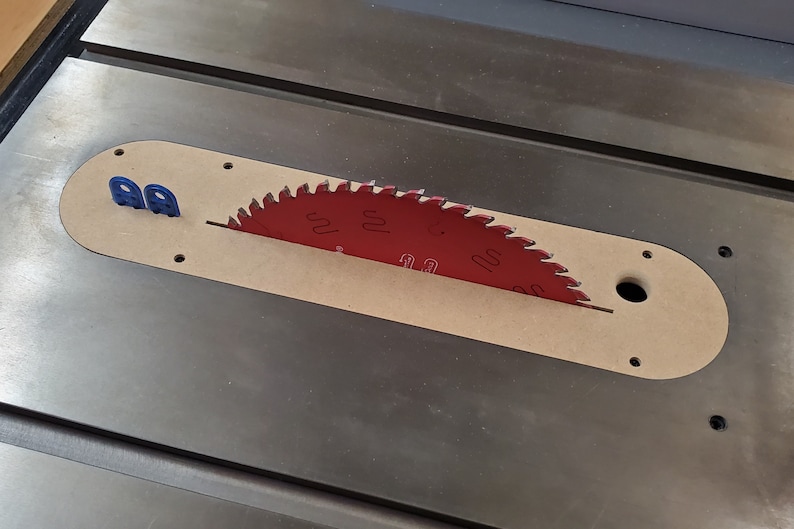 May include: A red saw blade with the number "100" printed on it is partially visible in a wooden slot on a metal table saw. The blade is surrounded by a wooden insert with a hole in the center. Two blue plastic pieces are visible on the left side of the insert.