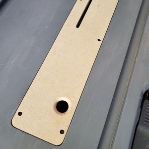 May include: A light brown wooden insert with a rectangular slot and a round hole. The insert is designed to fit into a larger surface, likely a table saw or workbench.