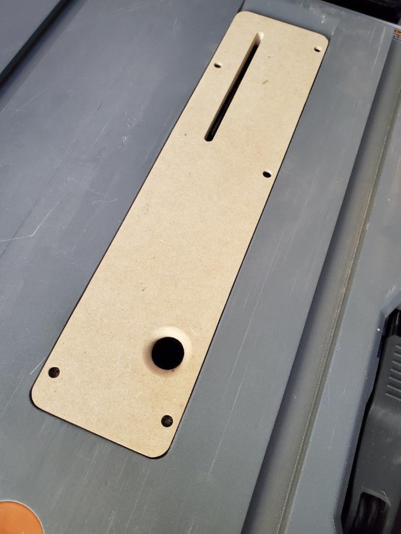 May include: A light brown wooden insert with a rectangular slot and a round hole. The insert is designed to fit into a larger surface, likely a table saw or workbench.
