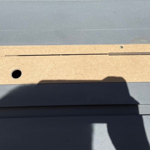 May include: A close-up of a light brown wooden panel with a long, thin slot cut into it. The panel is attached to a gray surface with screws. There is a small round hole in the panel.