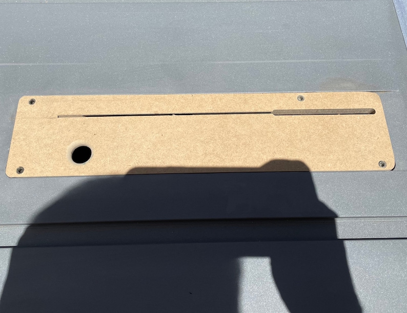 May include: A close-up of a light brown wooden panel with a long, thin slot cut into it. The panel is attached to a gray surface with screws. There is a small round hole in the panel.