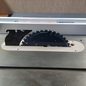 May include: A close-up of a table saw with a black saw blade and a silver metal fence. The saw blade is partially covered by a black plastic guard.