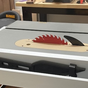 Puede incluir: Primer plano de una sierra de mesa con una hoja roja y una mesa de color gris. La hoja est&aacute; parcialmente cubierta por una protecci&oacute;n de madera. La sierra est&aacute; en un taller.