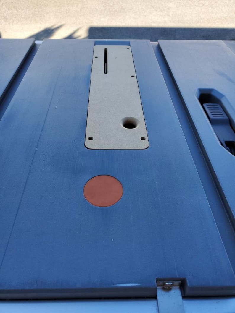May include: A close-up of a gray table saw with a light brown insert. The insert has a slot and a hole. There is a small, round, orange sticker on the table.