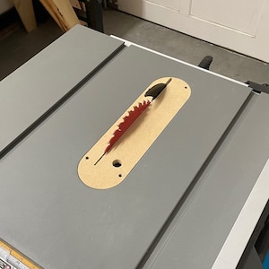Puede incluir: Primer plano de una sierra de mesa con un inserto de madera y una hoja de sierra roja. La hoja de sierra est&aacute; parcialmente insertada en el inserto.