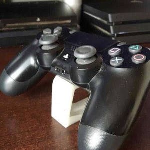 Op de afbeelding: Een zwarte PlayStation 4-controller ligt op een witte standaard. De controller heeft een zwart-wit ontwerp met knoppen en een touchpad.