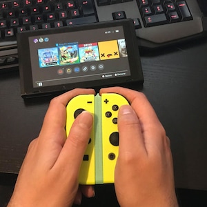May include: A person is holding a yellow and green Nintendo Switch Joy-Con controller. The controller is attached to a Nintendo Switch console, which is displaying the home screen with several game icons.